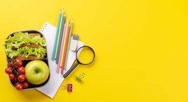 School supplies, stationery, and lunch box on yellow background. Education and nutrition. Flat lay with blank space