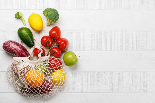 Mesh bag filled with a variety of vegetables. Flat lay over white wooden background with copy space