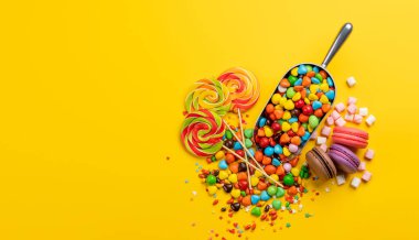 Çeşitli renkli şekerler, lolipoplar ve badem kurabiyeleri. Fotokopi alanı ile sarı arkaplan üzerinde düz yatıyordu