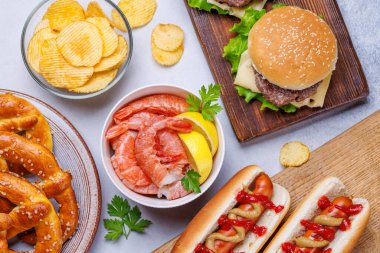 Canlı bir fotoğraf, çeşitli bira aperatifleri ve sulu bir çizburger, klasik bir sosisli sandviç, çıtır patates cipsi, tuzlu krakerler, taze karidesler, eğlenceli ve iştah açıcı bir sahne yaratmak da dahil olmak üzere fast food ürünleri sergiliyor.