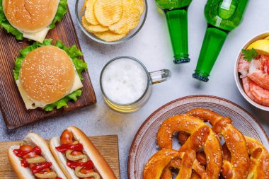 Canlı bir fotoğrafta çeşitli atıştırmalıklar ve fast food ürünleriyle çevrili bir bardak bira, sulu bir çizburger, klasik bir sosisli sandviç, çıtır patates cipsi, tuzlu krakerler, taze karidesler, eğlenceli ve iştah açıcı bir sahne yaratmak gibi.