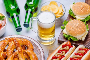 Canlı bir fotoğrafta çeşitli atıştırmalıklar ve fast food ürünleriyle çevrili bir bardak bira, sulu bir çizburger, klasik bir sosisli sandviç, çıtır patates cipsi, tuzlu krakerler, taze karidesler, eğlenceli ve iştah açıcı bir sahne yaratmak gibi.