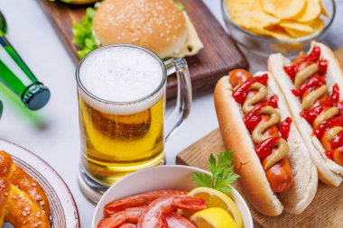 Canlı bir fotoğrafta çeşitli atıştırmalıklar ve fast food ürünleriyle çevrili bir bardak bira, sulu bir çizburger, klasik bir sosisli sandviç, çıtır patates cipsi, tuzlu krakerler, taze karidesler,