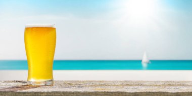 Bir bardak soğuk bira arka planda güzel bir deniz manzaralı bir masada oturur, plajda güneşli ve rahatlatıcı bir günün özünü yakalar.