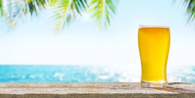 Bir bardak soğuk bira arka planda güzel bir deniz manzaralı bir masada oturur, plajda güneşli ve rahatlatıcı bir günün özünü yakalar.