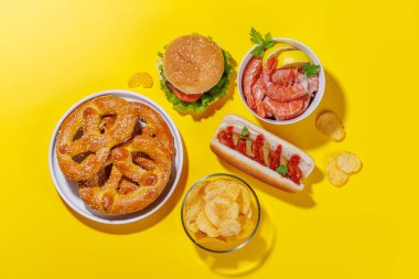 Canlı bir fotoğraf, çeşitli bira aperatifleri ve sulu bir çizburger, klasik bir sosisli sandviç, çıtır patates cipsi ve tuzlu krakerler, taze karidesler de dahil olmak üzere fast food ürünleri sergiliyor.