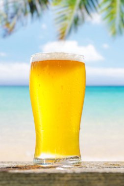 Bir bardak soğuk bira arka planda güzel bir deniz manzaralı bir masada oturur, plajda güneşli ve rahatlatıcı bir günün özünü yakalar.