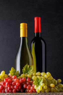 Üzümle eşleştirilmiş kırmızı ve beyaz şarap şişeleri, koyu bir zemin üzerine kurulmuş, zengin ve zarif bir ortam yaratıyor.