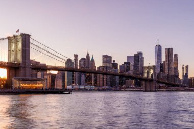 Manhattan 'a doğru uzanan Brooklyn Köprüsü' nün panoramik günbatımı manzarası, solan gökyüzüne karşı sıcak renklerle aydınlatılan gökdelenler.