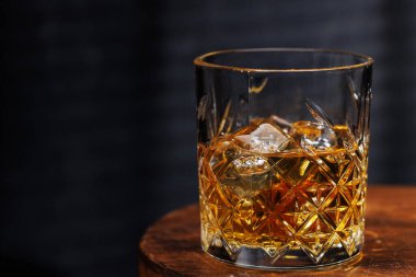 Koyu bir zemine dayanmış, ahşap bir masada dinlenen bir viski bardağı sıcak ve kırsal bir atmosferi çağrıştırıyor.
