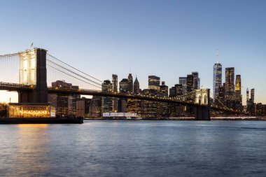 Manhattan 'a doğru uzanan Brooklyn Köprüsü' nün panoramik günbatımı manzarası, solan gökyüzüne karşı sıcak renklerle aydınlatılan gökdelenler.