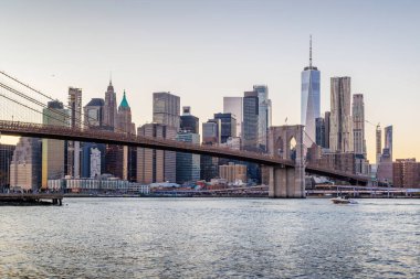 Manhattan 'a doğru uzanan Brooklyn Köprüsü' nün panoramik günbatımı manzarası, solan gökyüzüne karşı sıcak renklerle aydınlatılan gökdelenler.