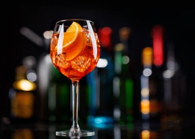 Bar masasında Aperol sprey kokteyli. Arka planda yanan şişelerin renkli bokeh 'i, dinamik ve canlı bir ortam yaratarak ortamı canlandırıyor.