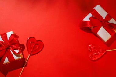 Kalp şeklinde bir lolipop romantik bir Sevgililer Günü hediye kutusuyla eşleştirilmiş, canlı kırmızı bir arka plana kurulmuş, aşkı ve kutlamayı simgeliyor.