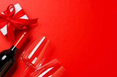 Bir kadeh ve bir şişe kırmızı şarap zarif bir sevgililer günü hediye kutusuyla, canlı kırmızı bir arka planda ayarlanmış, romantik ve lüks bir atmosfer yaratıyor.