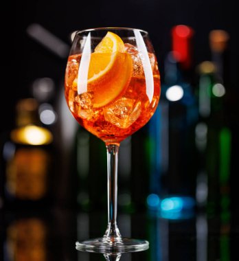 Bar masasında Aperol sprey kokteyli. Arka planda yanan şişelerin renkli bokeh 'i, dinamik ve canlı bir ortam yaratarak ortamı canlandırıyor.