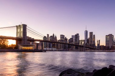 Manhattan 'a doğru uzanan Brooklyn Köprüsü' nün panoramik günbatımı manzarası, solan gökyüzüne karşı sıcak renklerle aydınlatılan gökdelenler.