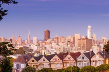 San Francisco 'daki Boyalı Kadınlar' ın canlı günbatımı manzarası. Renkli gökyüzünün ılık renkleri altında parlayan ikonik Viktorya dönemi evleri. Arka planda gökyüzü var.