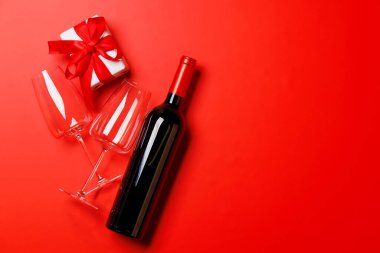 Bir kadeh ve bir şişe kırmızı şarap zarif bir sevgililer günü hediye kutusuyla, canlı kırmızı bir arka planda ayarlanmış, romantik ve lüks bir atmosfer yaratıyor.