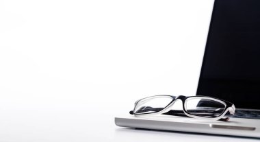 Pürüzsüz bir dizüstü bilgisayar ve temiz beyaz bir yüzey üzerinde okuma gözlüklerinin bulunduğu bir ofis masasının yakın çekimi, minimalist ve profesyonel bir titreşim için yüksek anahtarlı aydınlatma ile sunulmaktadır.