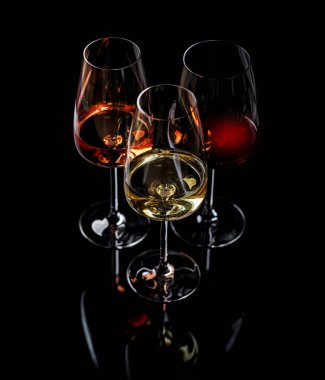 Kırmızı, beyaz ve gül şarabıyla dolu üç şarap kadehi siyah arka planda zarifçe duruyor. Şarabın çeşitliliğinin ve zarafetinin canlı bir gösterisi.