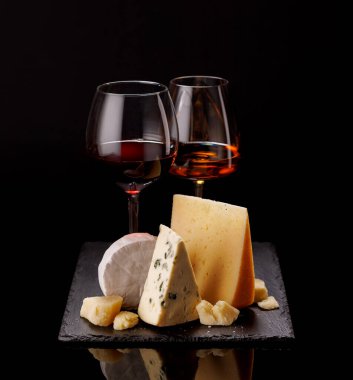 Dramatik bir karanlık arkaplan üzerine kurulmuş, bir bardak şarapla birlikte, bir tahtada sunulan rafine edilmiş peynir seçimi. Gurme hoşgörüsü için mükemmel bir ortam.