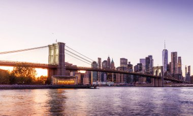 Manhattan 'a doğru uzanan Brooklyn Köprüsü' nün panoramik günbatımı manzarası, solan gökyüzüne karşı sıcak renklerle aydınlatılan gökdelenler.