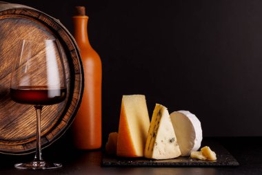 Dramatik bir karanlık arkaplan üzerine kurulmuş, bir bardak şarapla birlikte, bir tahtada sunulan rafine edilmiş peynir seçimi. Gurme hoşgörüsü için mükemmel bir ortam.