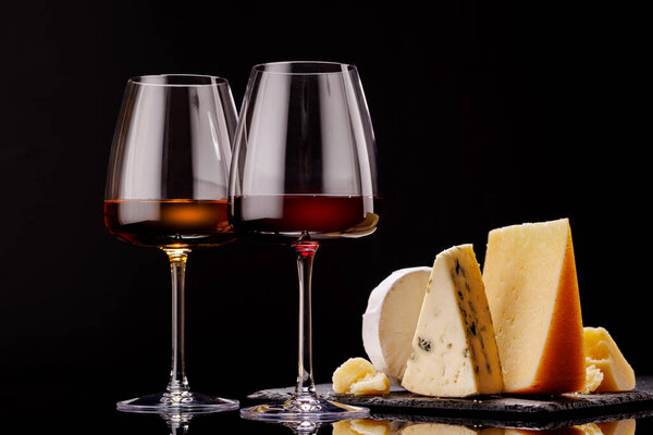 A refined selection of aged cheeses presented on a board, accompanied by a glasses of wine, set against a dramatic dark background. A perfect setting for a gourmet indulgence