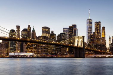 Manhattan 'a doğru uzanan Brooklyn Köprüsü' nün panoramik günbatımı manzarası, solan gökyüzüne karşı sıcak renklerle aydınlatılan gökdelenler.