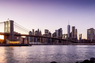 Manhattan 'a doğru uzanan Brooklyn Köprüsü' nün panoramik günbatımı manzarası, solan gökyüzüne karşı sıcak renklerle aydınlatılan gökdelenler.