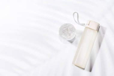 Parlak beyaz bir arka plana yerleştirilmiş parlak bir su şişesi, doğal, güneşli gölgelerle vurgulanmış, tazeliği ve modern minimalizmi aktarıyor.