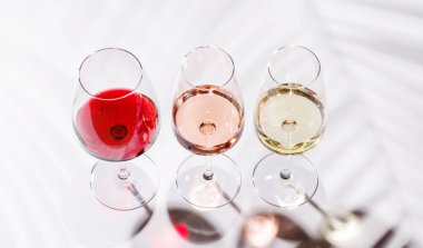 Kırmızı, beyaz ve gül şarabıyla dolu üç şarap kadehi parlak, minimalist arka planda zarif bir şekilde duruyor. Şarabın çeşitliliğinin ve zarafetinin canlı bir gösterisi.