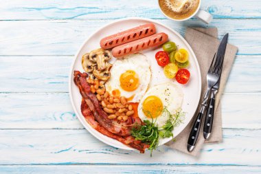 Günün başlangıcında sıcak bir fincan kahveyle sunulan, güneşli, kızarmış pastırmalı, sosisli ve taze domatesli klasik bir kahvaltı tabağı.