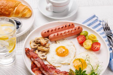 Günün başlangıcında sıcak bir fincan kahveyle sunulan, güneşli, kızarmış pastırmalı, sosisli ve taze domatesli klasik bir kahvaltı tabağı.