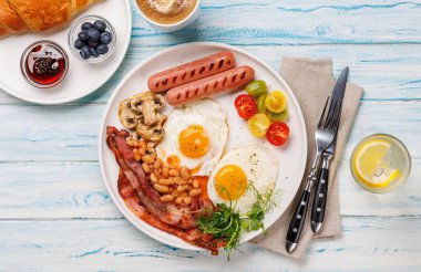 Günün başlangıcında sıcak bir fincan kahveyle sunulan, güneşli, kızarmış pastırmalı, sosisli ve taze domatesli klasik bir kahvaltı tabağı.