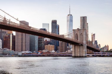 Manhattan 'a doğru uzanan Brooklyn Köprüsü' nün panoramik günbatımı manzarası, solan gökyüzüne karşı sıcak renklerle aydınlatılan gökdelenler.