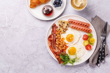 Günün başlangıcında sıcak bir fincan kahveyle sunulan, güneşli, kızarmış pastırmalı, sosisli ve taze domatesli klasik bir kahvaltı tabağı.