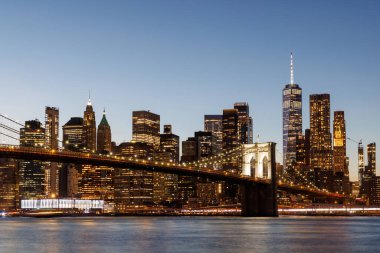 Manhattan 'a doğru uzanan Brooklyn Köprüsü' nün panoramik günbatımı manzarası, solan gökyüzüne karşı sıcak renklerle aydınlatılan gökdelenler.