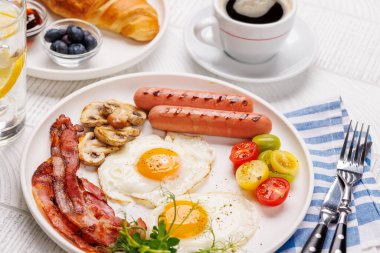Günün başlangıcında sıcak bir fincan kahveyle sunulan, güneşli, kızarmış pastırmalı, sosisli ve taze domatesli klasik bir kahvaltı tabağı.