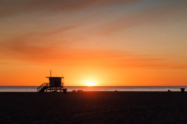 Gün batımında Los Angeles plajında, cankurtaran kulesi silueti parlayan turuncu ve mor bir ufuk çizgisi, hafif dalgalar ve sakin bir ortam.