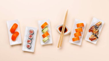 Çeşit çeşit suşi ve maki börekleri, taze malzemeler ve canlı renkler içeren tabaklar üzerinde güzelce düzenlenmiş, bej arkaplanda düz bir şekilde yatıyordu.