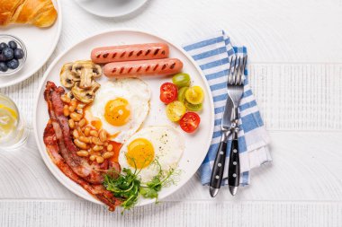 Günün başlangıcında sıcak bir fincan kahveyle sunulan, güneşli, kızarmış pastırmalı, sosisli ve taze domatesli klasik bir kahvaltı tabağı.