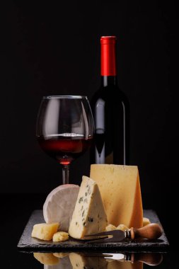 Dramatik bir karanlık arkaplan üzerine kurulmuş, bir bardak şarapla birlikte, bir tahtada sunulan rafine edilmiş peynir seçimi. Gurme hoşgörüsü için mükemmel bir ortam.