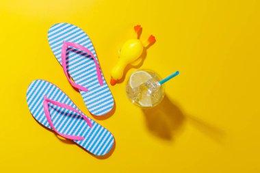 Parlak bir arka planda parmak arası terlikleri ve kokteyli olan canlı bir yaz tatili sahnesi, kopya uzayı ile eğlenceli ve tropikal bir atmosferi çağrıştırıyor.