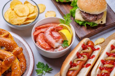 Canlı bir fotoğraf, çeşitli bira aperatifleri ve sulu bir çizburger, klasik bir sosisli sandviç, çıtır patates cipsi ve tuzlu krakerler, taze karidesler de dahil olmak üzere fast food ürünleri sergiliyor.