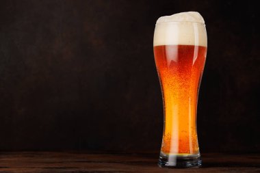 Ahşap bir masanın üzerinde köpüklü bir kafayla bir bardak kehribar birası. Bira, bar ve içecek sunumları için ferahlatıcı bir konsept