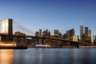 Manhattan 'a doğru uzanan Brooklyn Köprüsü' nün panoramik günbatımı manzarası, solan gökyüzüne karşı sıcak renklerle aydınlatılan gökdelenler.
