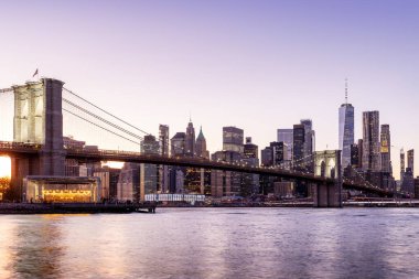 Manhattan 'a doğru uzanan Brooklyn Köprüsü' nün panoramik günbatımı manzarası, solan gökyüzüne karşı sıcak renklerle aydınlatılan gökdelenler.