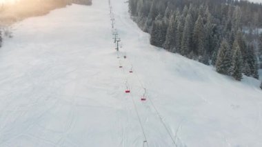 Kış dağları, gün batımında kayak pistleri ve kayak sandalyeleriyle yükseliyor. Boş dağ kayak pistinin hava aracı görüntüsü, karla kaplı ladin ormanı ve kar yağışı sırasında teleferik. Yörünge atışı